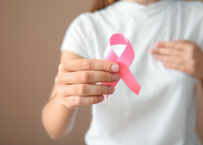 Frau in weißem T-Shirt hält sich mit einer Hand an der Brust und in der anderen ein rosa Schleife